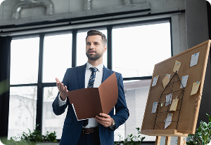Smiling businessman in suit presents, gesturing with a folder in hand. Behind him, a corkboard with a project plan and a specific Form ID